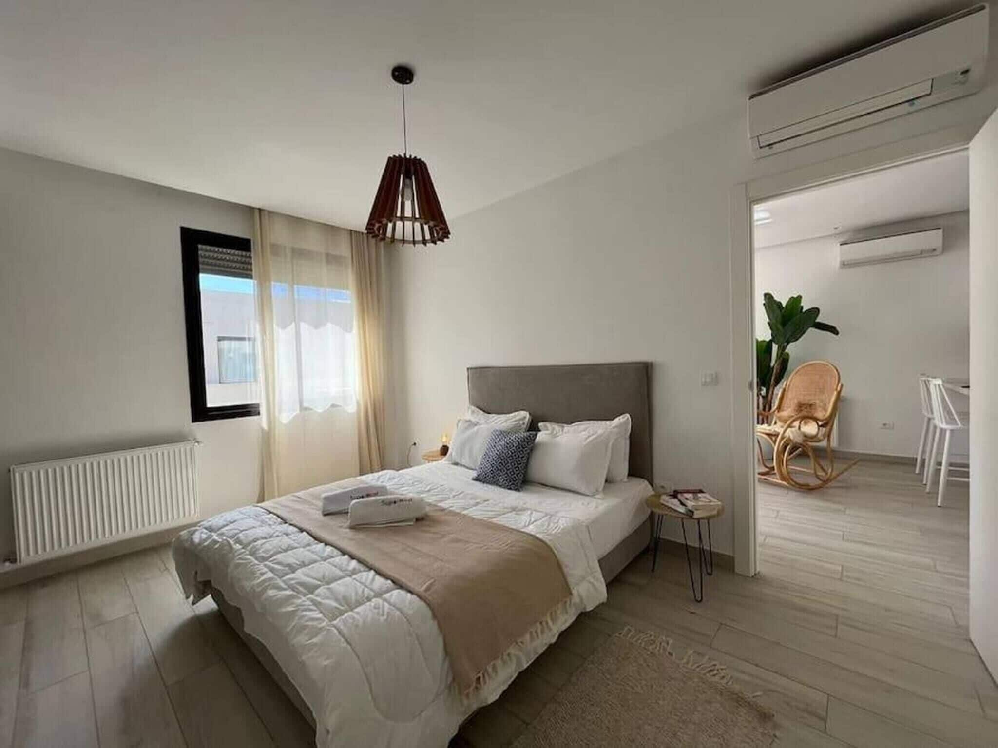 A modern bedroom with a large bed, a bedside table, a hanging light fixture, and a view of the outside through a window.
