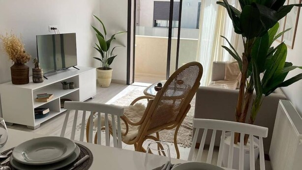 Un comedor con una mesa blanca para dos personas, un televisor sobre un soporte blanco y plantas en macetas.