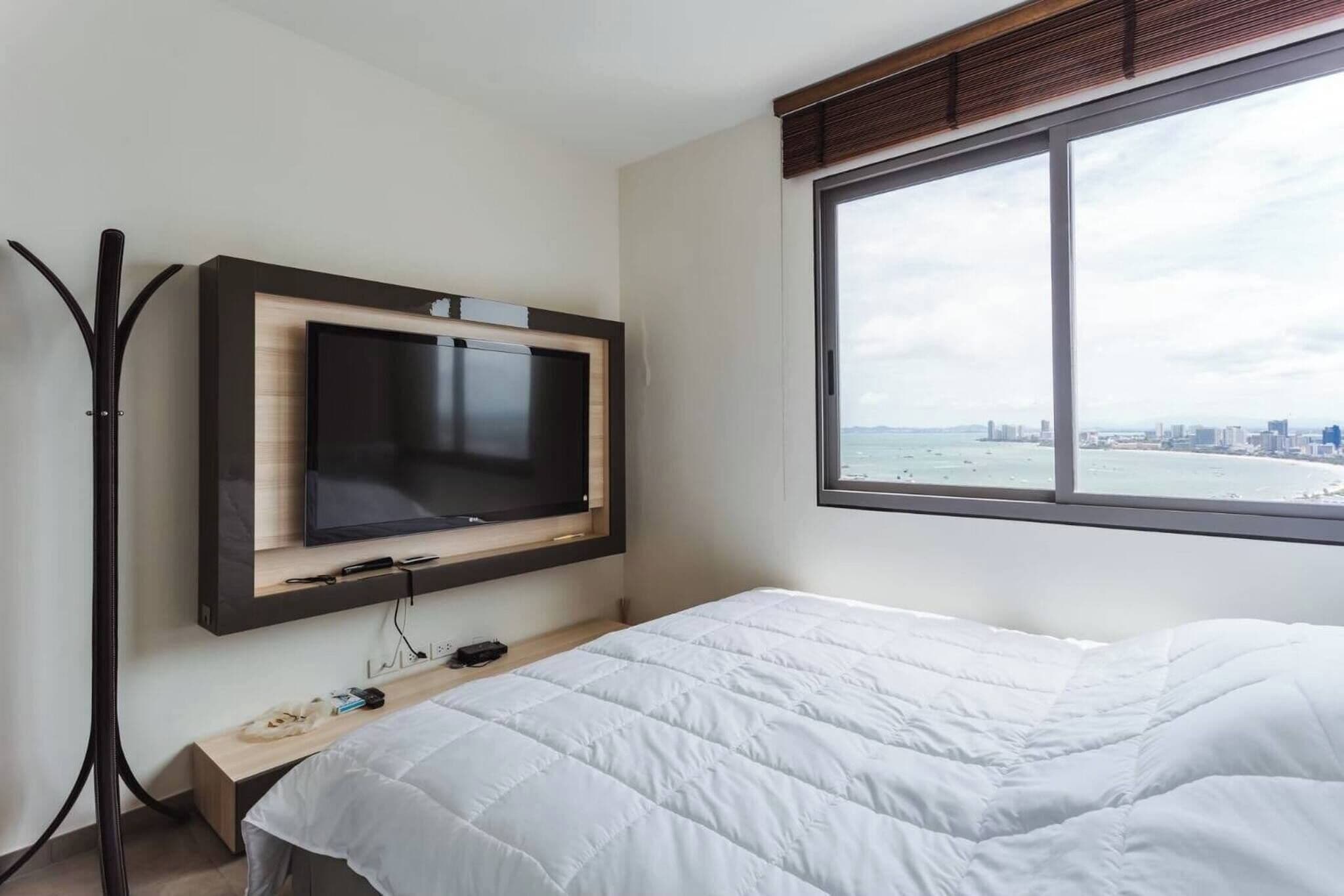 A hotel room with a bed, a TV mounted on the wall, a window with a city view, and a desk with a chair.