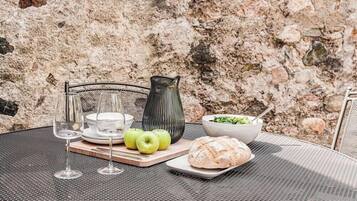 Mesa al aire libre con ensalada en un bol, hogaza de pan y tres manzanas, copas de vino y un florero sobre una mesa de metal.