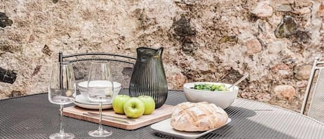 A table set with a bowl of salad, a loaf of bread, and three apples, with wine glasses and a vase on a metal table outdoors.