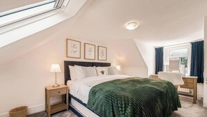 A bedroom with a sloped ceiling, a bed with a green blanket, two bedside tables with lamps, a window with curtains, and framed pictures on the wall. - Cromar Cottage in Aberdeenshire (Ballater)