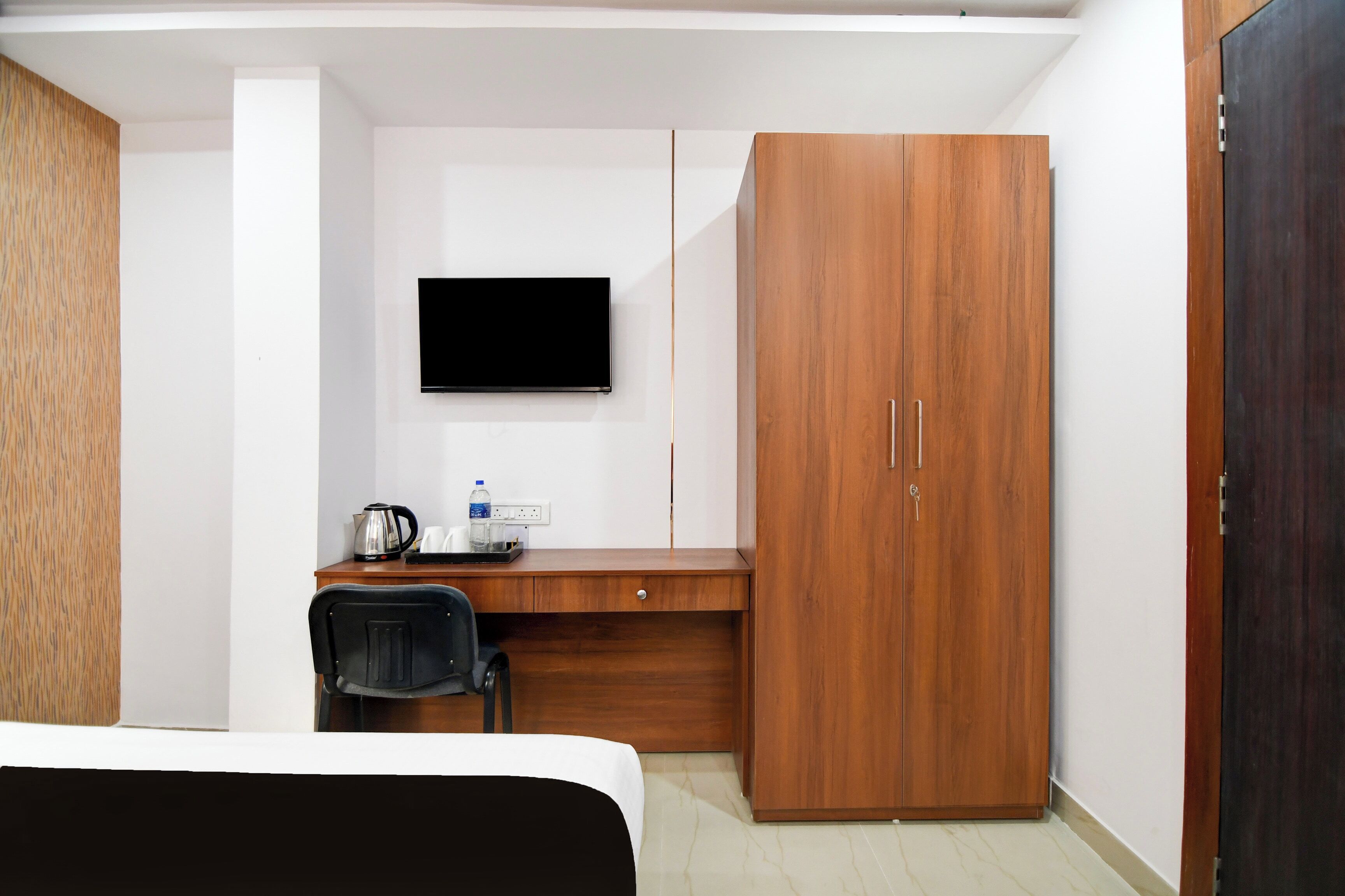 A hotel room with a bed, a desk with a chair, a wardrobe, and a flat-screen TV mounted on the wall.