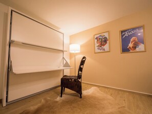 Une pièce avec un écran blanc rétractable, une chaise à motifs évoquant la fourrure, un lampadaire et deux affiches encadrées accrochées au mur.