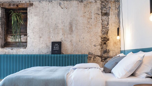 Un dormitorio con pared de piedra, una planta colgante, un cartel, una cama con ropa de cama blanca y un cabecero azul.