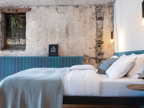 A bedroom with a stone wall, a hanging plant, a sign, a bed with white bedding, and a blue headboard.