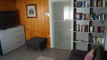 Ein Wohnzimmer mit Fernseher, Bücherregal, einer Couch und einer Glastür zum Balkon.