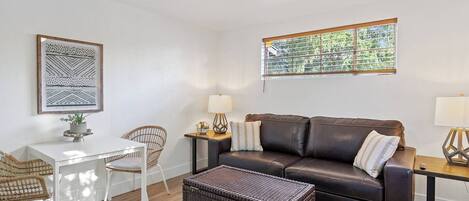 A living room with a brown leather sofa, a white table, wicker chairs, a ceiling fan, and a window with a view of trees.