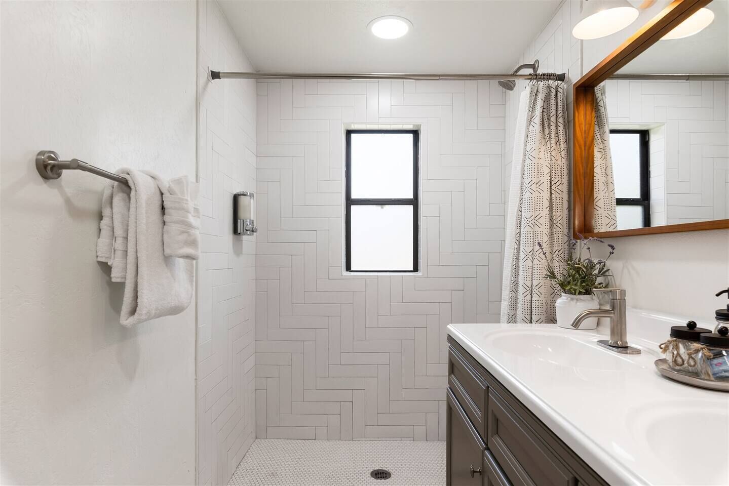 A modern bathroom with a herringbone tile pattern, a shower with a glass door, a vanity with a mirror, and a window providing natural light.