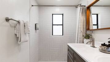 A modern bathroom with a herringbone tile pattern, a shower with a glass door, a vanity with a mirror, and a window providing natural light.