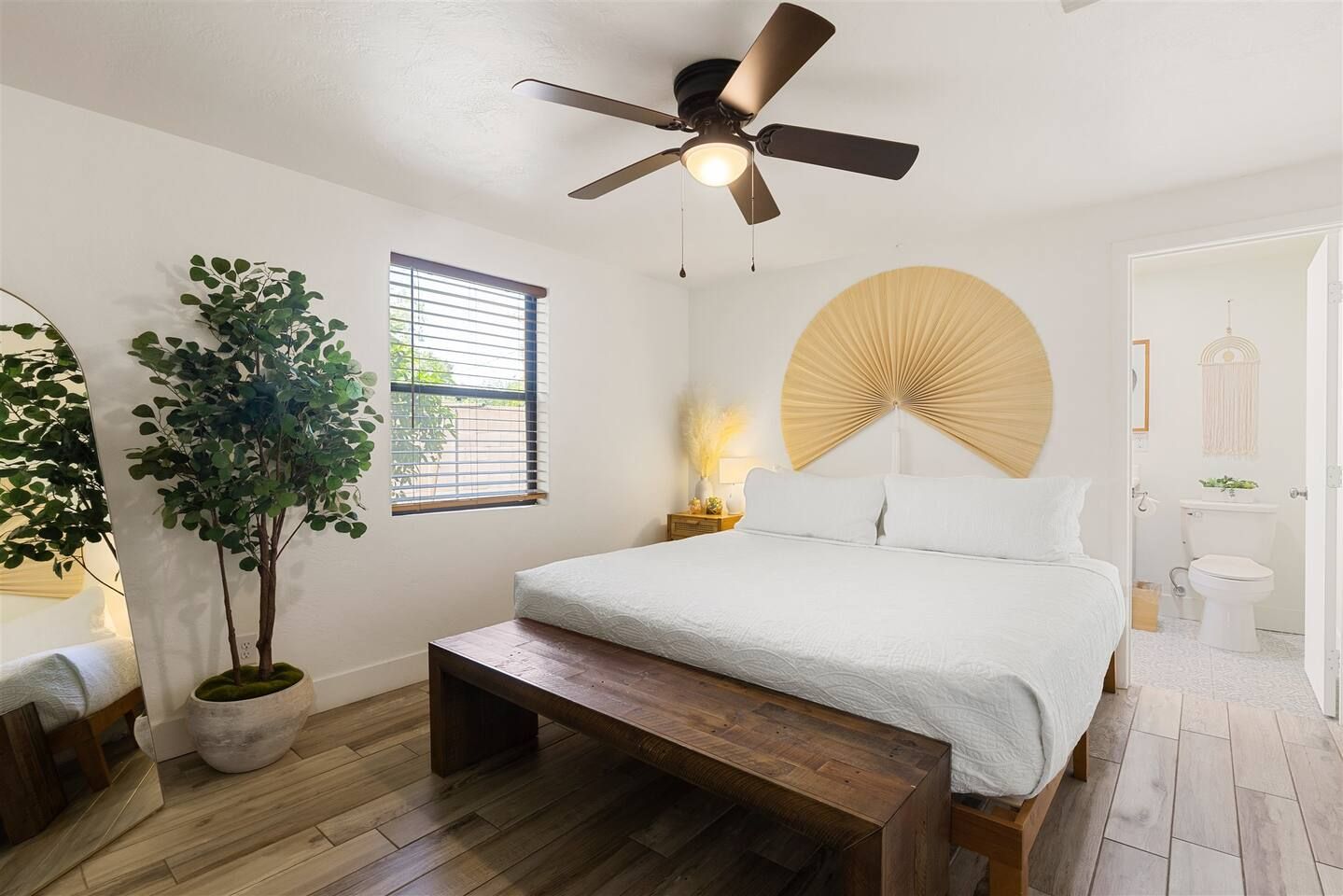 A bedroom with a bed, a ceiling fan, a window, a bedside table, a nightstand, a potted plant, and a door leading to a bathroom.