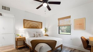 Un dormitorio con cama, mesitas de noche, ventilador de techo, ventana con vistas y un cartel en la pared.