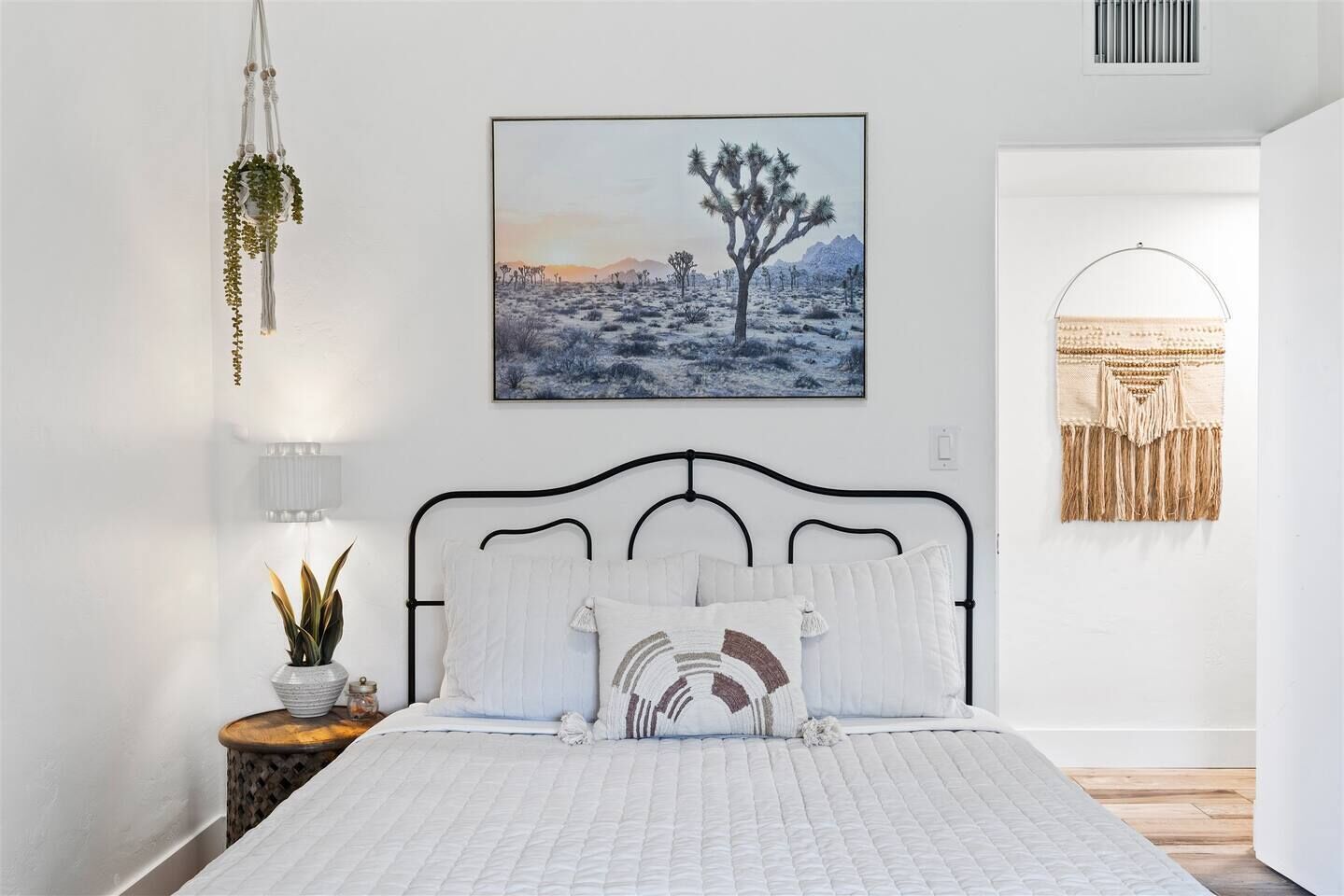 Une chambre à coucher avec un lit, une plante en macramé suspendue, un tableau représentant un paysage désertique encadré et une décoration murale en macramé.