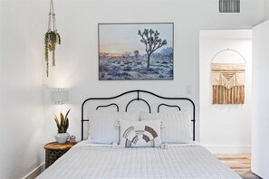 A bedroom with a bed, a hanging macrame plant, a framed desert landscape painting, and a macrame wall hanging.