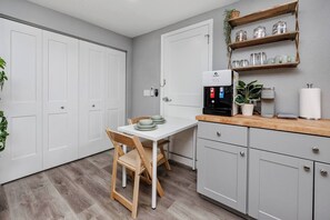 A compact kitchen with a coffee machine, a dining table with chairs, and a shelf with jars.