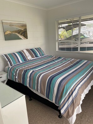 A bedroom with a bed, a nightstand, a window with blinds, and a wall-mounted picture. - Whitianga downstairs flat with car & boat parking close to beach (Whitianga)