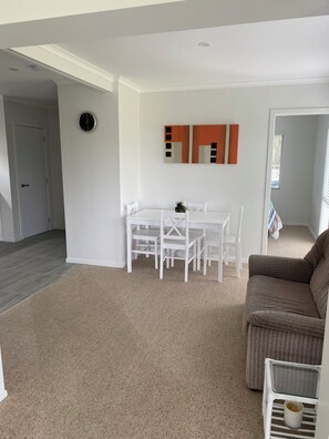 A living room with a brown sofa, a white coffee table, and a dining area with a white table and chairs. - Whitianga downstairs flat with car & boat parking close to beach (Whitianga)