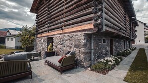 Eine Blockhütte mit Steinfundament, Sitzbereich im Freien mit Stühlen und einer Couch sowie ein gepflegter Garten.