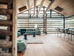 A log cabin interior with a wooden ceiling, a green sofa set, a dining table with chairs, and a large window.