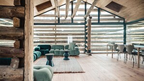 Ein Blockhaus-Interieur mit Holzbalkendecke, einem grünen Sofasitzgruppe, einem Esstisch mit Stühlen und einem großen Fenster, das für natürliches Licht sorgt.