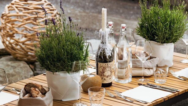 Une table en bois, dressée pour le dîner, agrémentée de plantes en pot, d’une bouteille de vin et de verres.