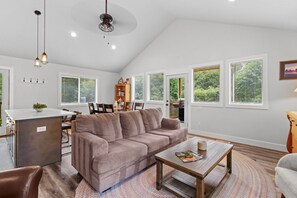 A spacious living room with a brown sofa, a wooden coffee table, and a kitchen area in the background. - 14-Acre Mountain Escape - Hot Tub + Fire Pit (Nehalem)