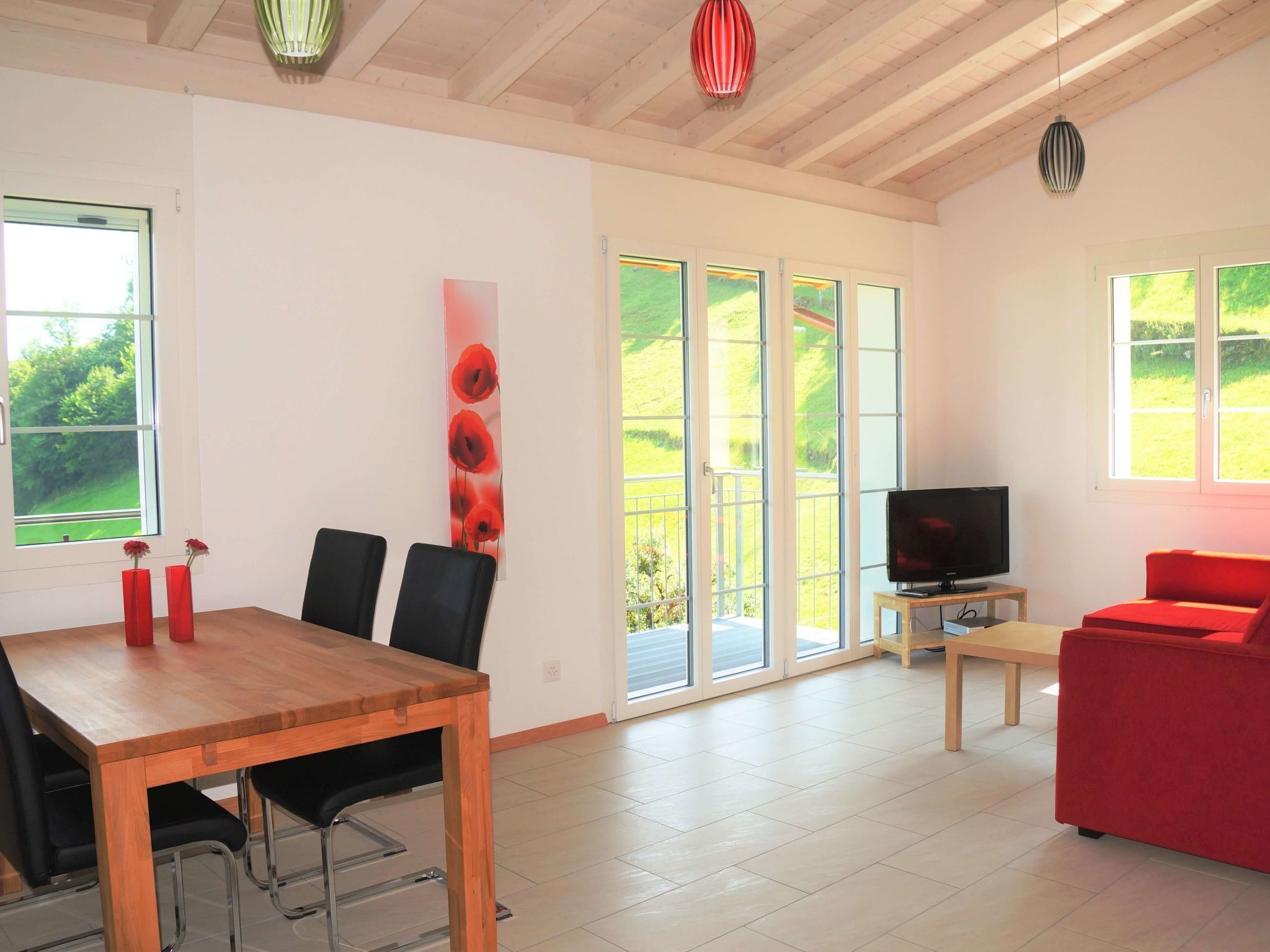 Ein modernes Wohnzimmer mit Esstisch aus Holz, schwarzen Stühlen, einem roten Sofa, einem Fernseher auf einem Holzboden und großen Fenstern mit Blick auf Grünflächen.
