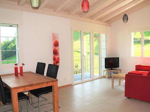 Un salon moderne avec une table à manger en bois, des chaises noires, un canapé rouge, un téléviseur posé sur un meuble en bois et de grandes fenêtres donnant sur un paysage verdoyant.
