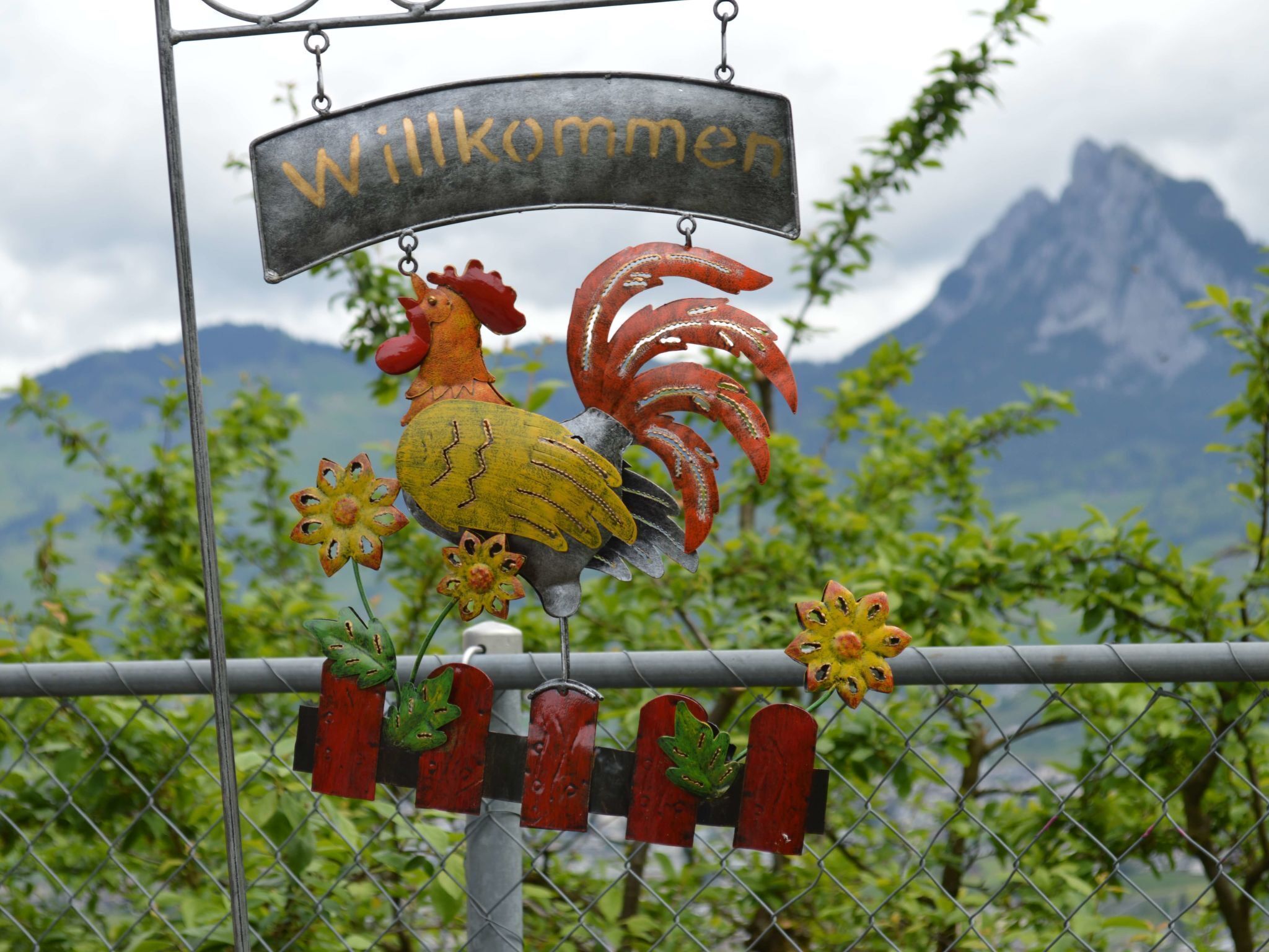 A metal sign with a rooster and flowers, with a mountainous landscape in the background.