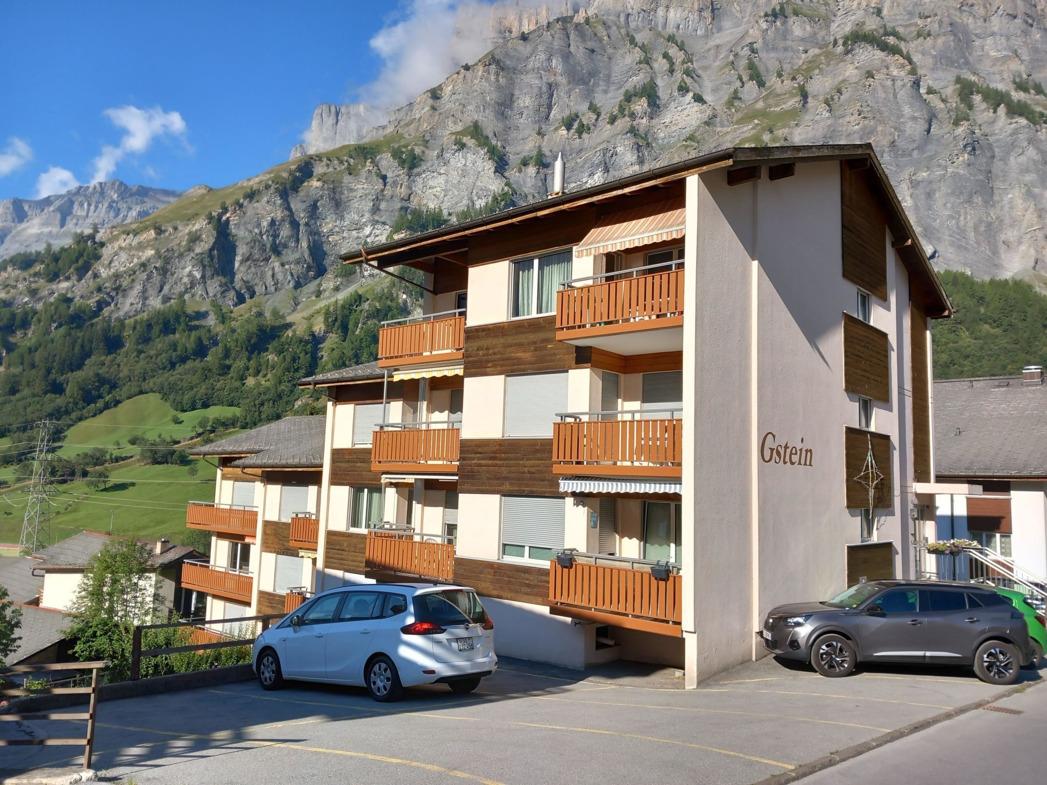 A multi-story hotel with balconies, a parking area, and a mountainous backdrop.