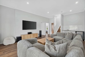 A modern living room with a grey sectional sofa, a wooden coffee table, and a flat-screen TV mounted on the wall. - Beachside Family Escape 92 Newell Avenue Middleton Spacious 4-Bedroom Holiday Home Sleeps 10 one street from Middleton Beach (Middleton)