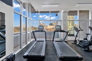 Ein Fitnessraum mit Laufbändern und einem Crosstrainer, große Fenster mit Blick auf die Stadt und ein Schild, das eine 24-Stunden-Videoüberwachung anzeigt.
