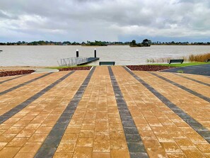 A paved walkway leading to a body of water with a bench and a small island visible in the distance. - Stillwater - holiday home with a private jetty (Wellington East)