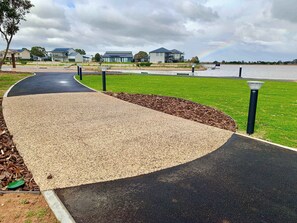 A paved walkway with a curved design, bordered by grass and mulch, leading to a body of water with houses in the background.