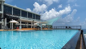 Uma piscina com superfície de água azul cristalina, rodeada por espreguiçadeiras e guarda-sóis, e um edifício com grandes janelas e varandas ao fundo.
