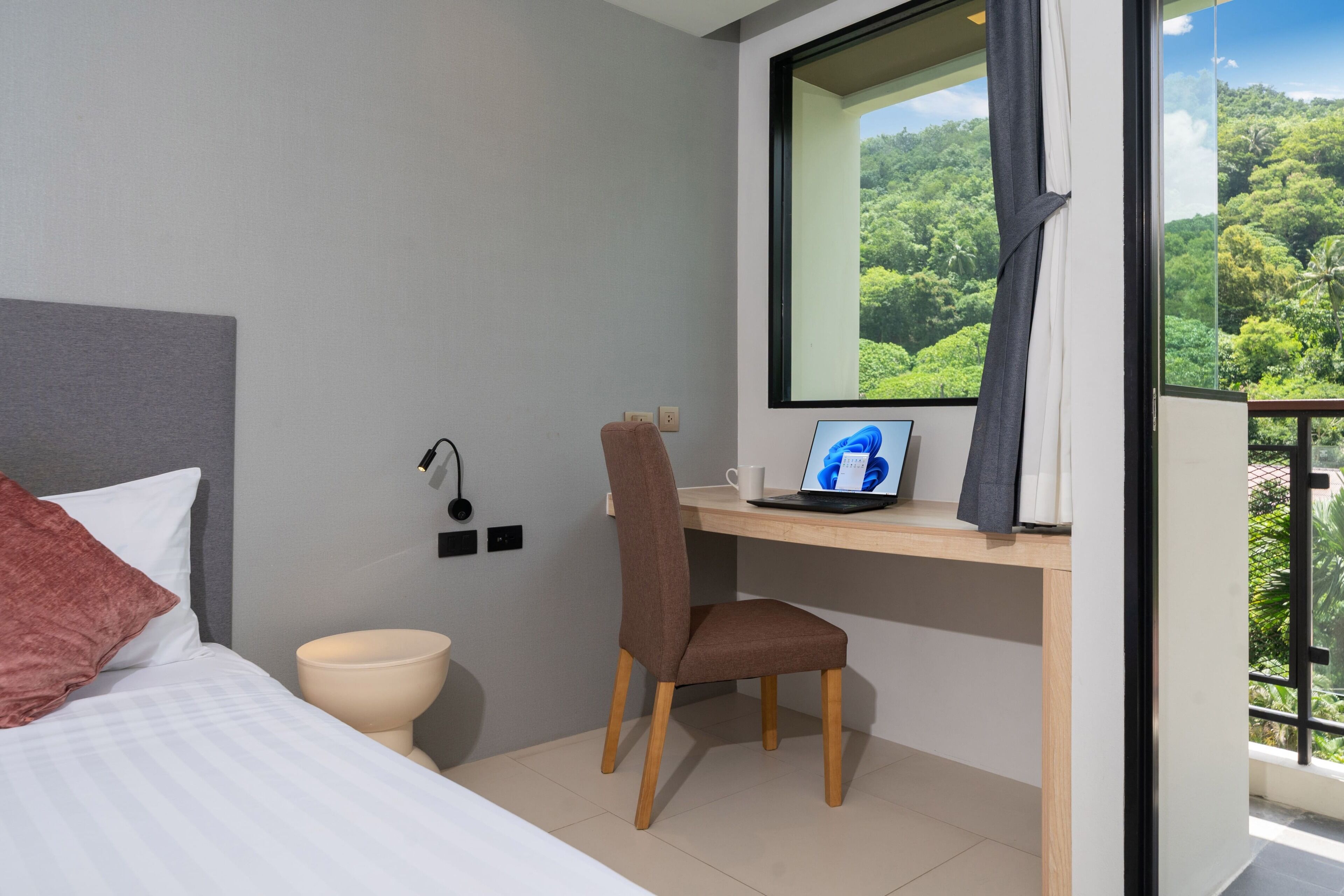 Camera d'albergo con un letto, una scrivania con un portatile, una sedia e vista su una foresta attraverso una finestra.