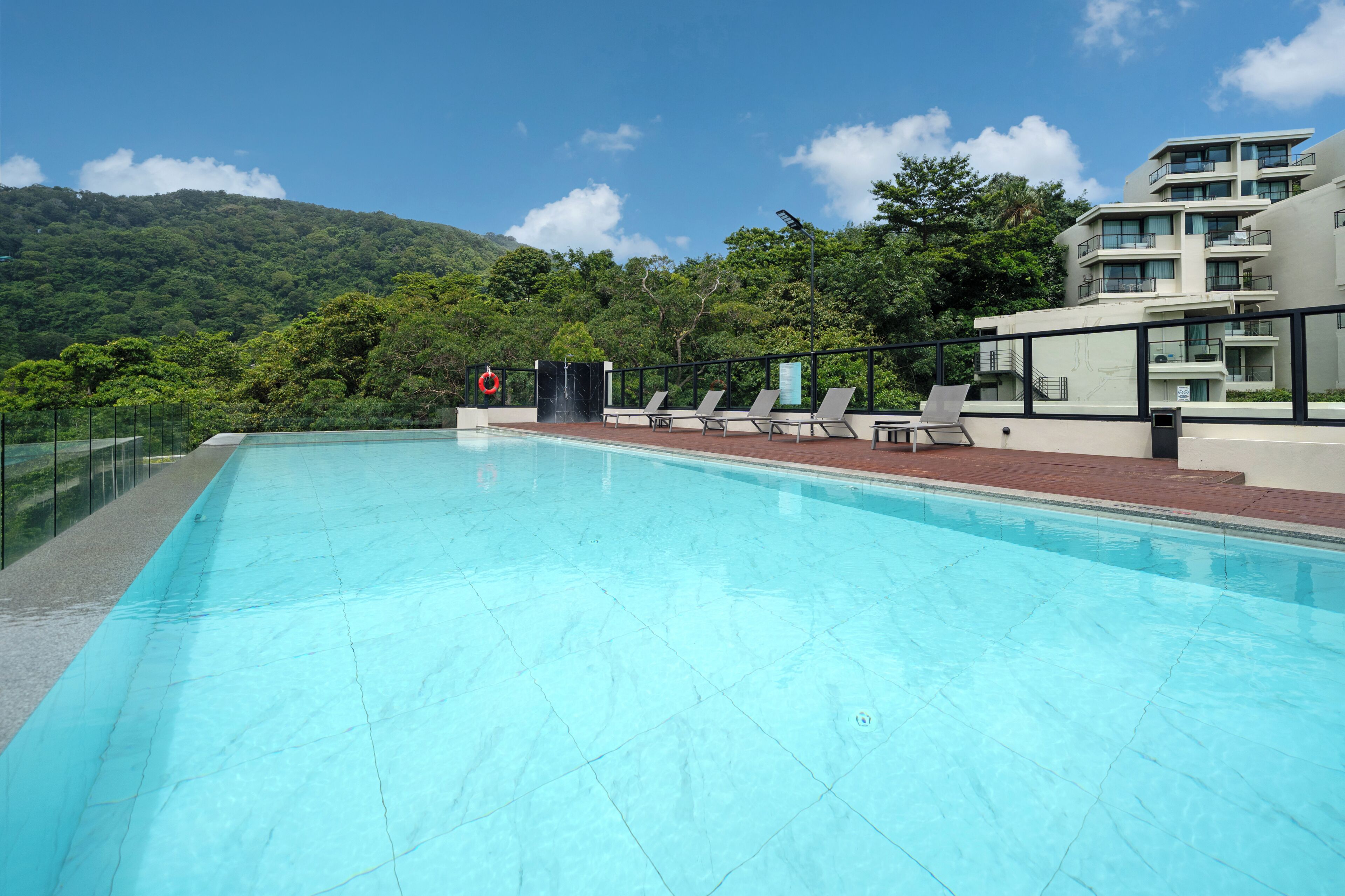 A clear blue swimming pool with lounge chairs and a safety sign, surrounded by lush greenery and a modern building.