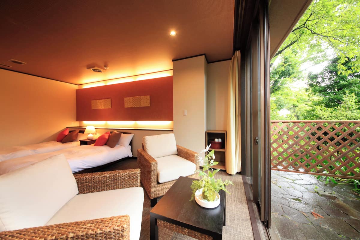 A hotel room with a bed, two sofas, a small table with a plant, and a view of greenery through a window.