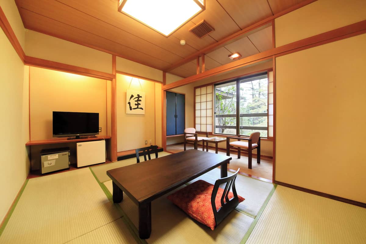 A traditional Japanese room with tatami flooring, a low table, chairs, a television, and a sliding door leading to a window with a view of trees.