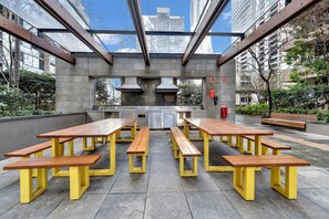 An outdoor dining area with multiple wooden tables and benches, a modern kitchen with stainless steel appliances, and a glass ceiling allowing natural light. - High-Rise 1-Bed Apartment Near Southern Cross (Melbourne)