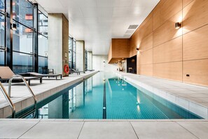 An indoor swimming pool with clear blue water, a lifebuoy, and a modern design with glass walls and wooden paneling. - High-Rise 1-Bed Apartment Near Southern Cross (Melbourne)