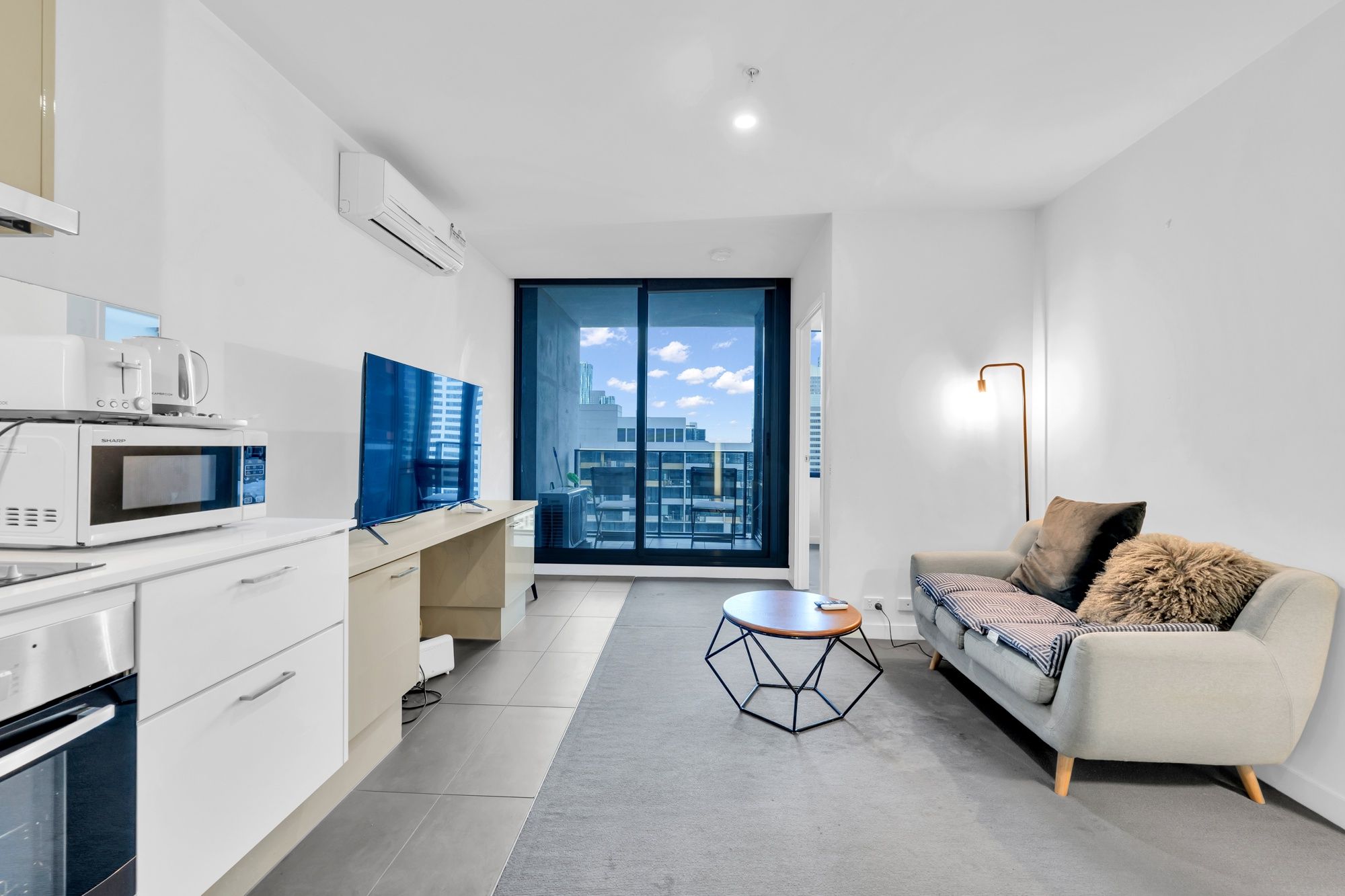 A modern apartment with a kitchenette, a sofa, a coffee table, and a view of the city through large windows.