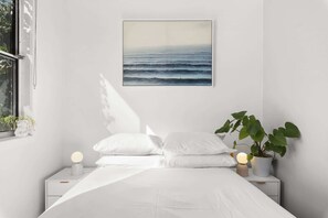 A bedroom with a bed, bedside tables, a potted plant, and a wall-mounted picture of the ocean.