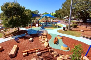 Ein Spielplatz mit bunten Spielgeräten, einem schattigen Bereich und einer naturnahen Spielzone mit Steinen und Baumstämmen.