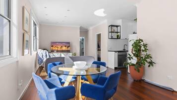 A modern living room with a round glass table, blue chairs, a painting of a sunset, and a view into the kitchen area.