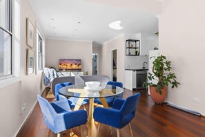 Ein modernes Wohnzimmer mit einem runden Glastisch, blauen Stühlen, einem Sonnenuntergangsgemälde und Blick in den Küchenbereich.