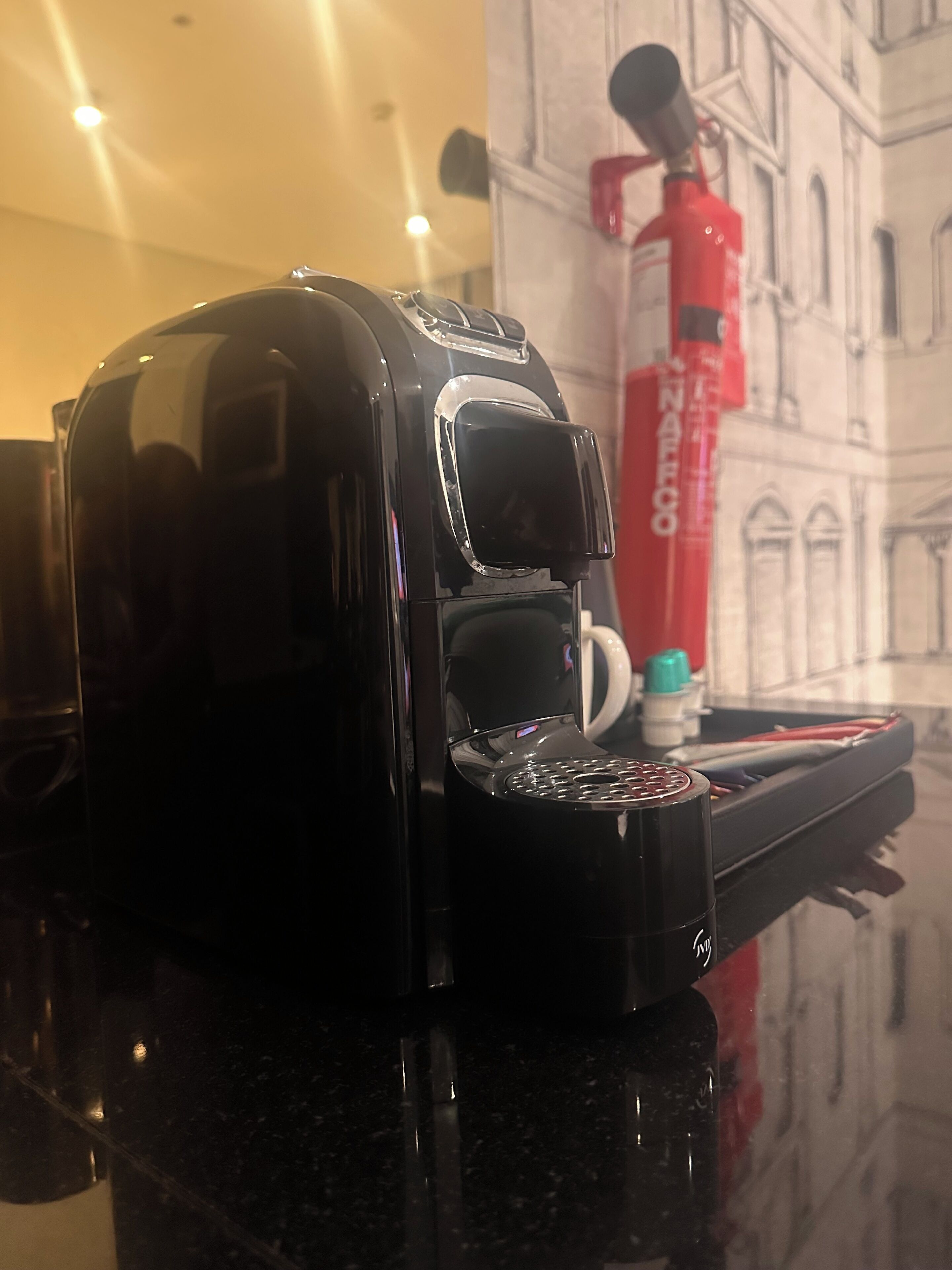A black coffee maker on a countertop with a red fire extinguisher in the background.