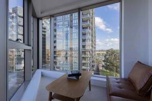 A modern living room with a sofa, a coffee table, and a view of high-rise buildings through large windows. - Skyline-View 2-Bed Apartment Near Flemington (Flemington)