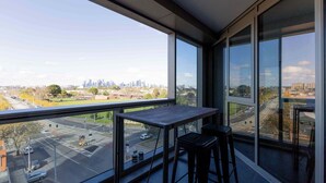A balcony with a view of a city skyline, a table, and two bar stools. - Skyline-View 2-Bed Apartment Near Flemington (Flemington)