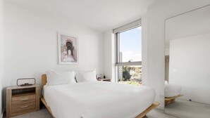 A bedroom with a large bed, a bedside table, a nightstand, a window with a view, and a framed picture on the wall. - Skyline-View 2-Bed Apartment Near Flemington (Flemington)